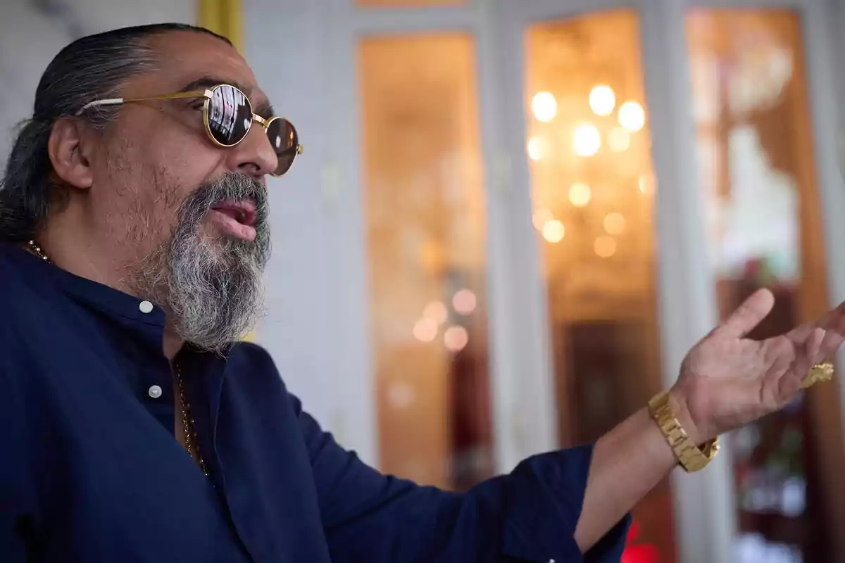Hombre de cabello largo y barba canosa usando gafas de sol y camisa azul gesticulando con la mano en un ambiente interior elegante con luces cálidas de fondo Hombre de cabello largo y barba canosa usando gafas de sol y camisa azul gesticulando con la mano en un ambiente interior elegante con luces cálidas de fondo