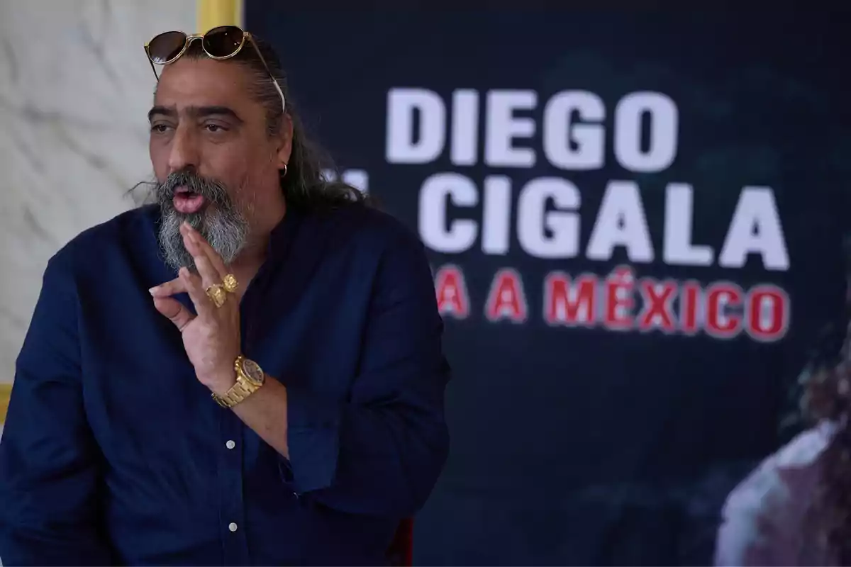 Hombre de cabello largo y barba canosa con gafas en la cabeza y camisa azul gesticulando con la mano frente a un cartel que dice Diego El Cigala va a México Hombre de cabello largo y barba canosa con gafas en la cabeza y camisa azul gesticulando con la mano frente a un cartel que dice Diego El Cigala va a México