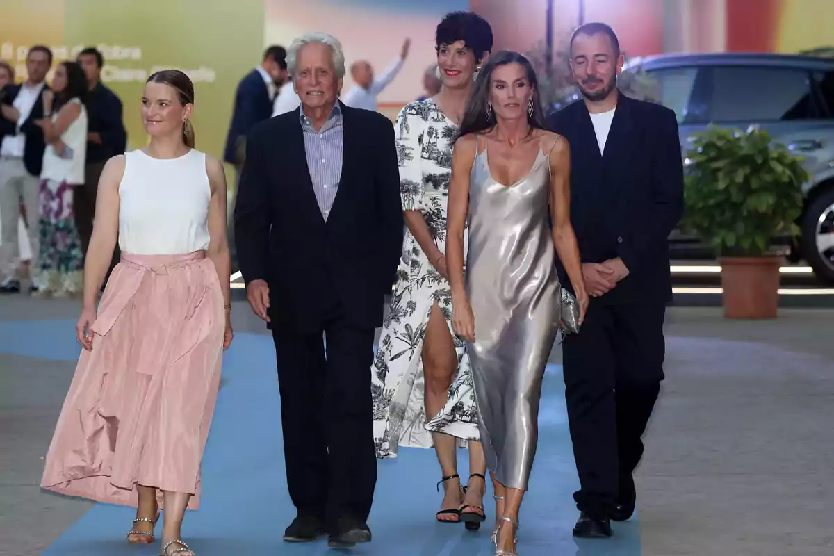 Cinco personas elegantemente vestidas caminan juntas por una alfombra azul en un evento social al aire libre