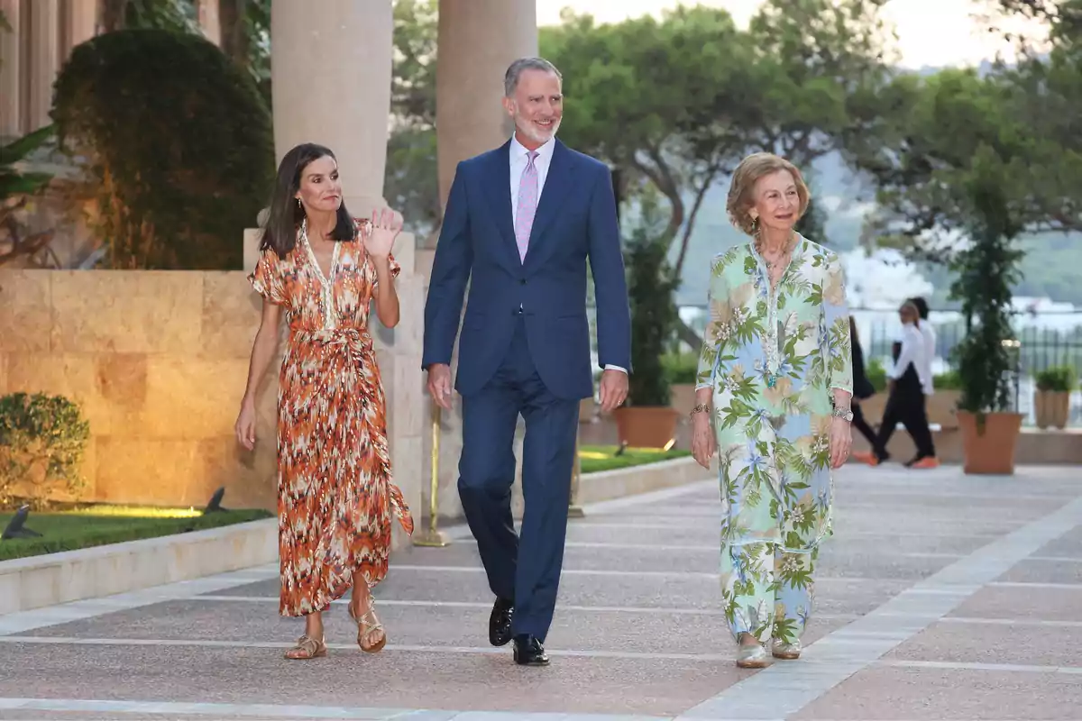 Tres personas elegantemente vestidas caminan al aire libre por un camino rodeado de vegetación y columnas, una de ellas saluda con la mano. Tres personas elegantemente vestidas caminan al aire libre por un camino rodeado de vegetación y columnas, una de ellas saluda con la mano.