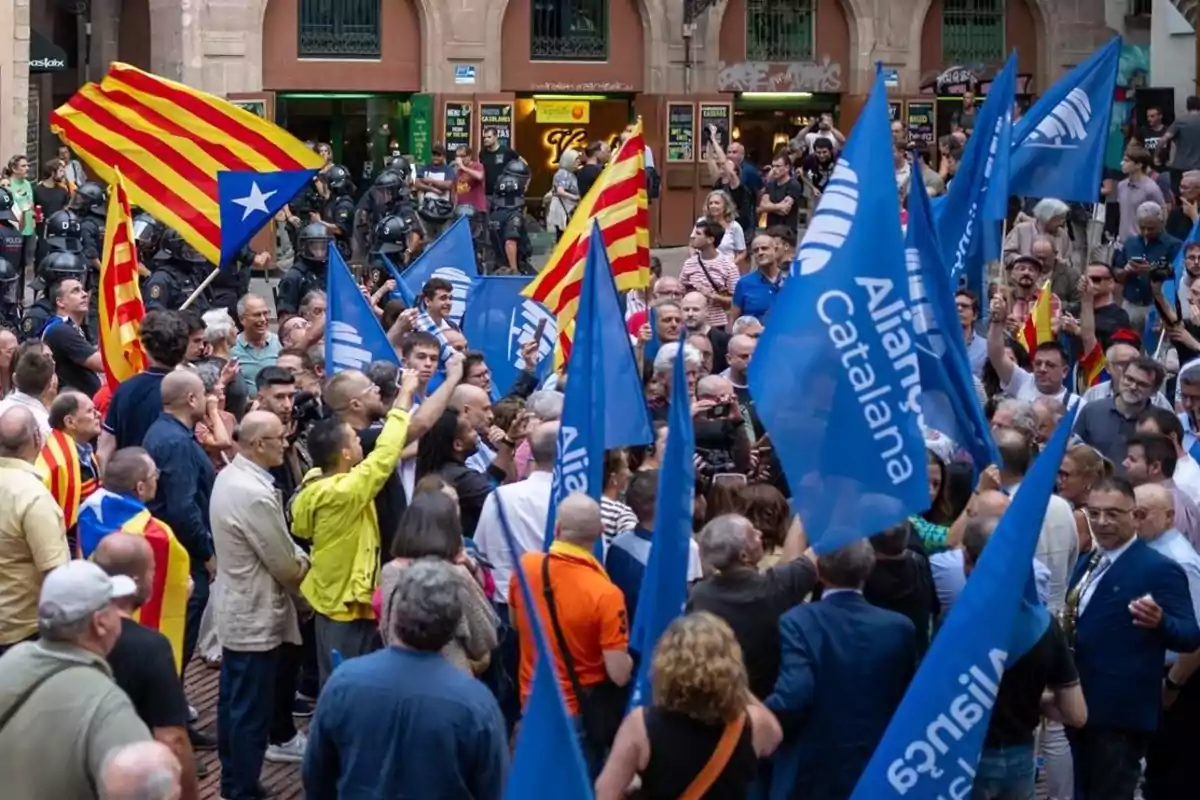 Manifestación con personas portando banderas esteladas y banderas azules de Aliança Catalana en una plaza, con presencia policial al fondo.