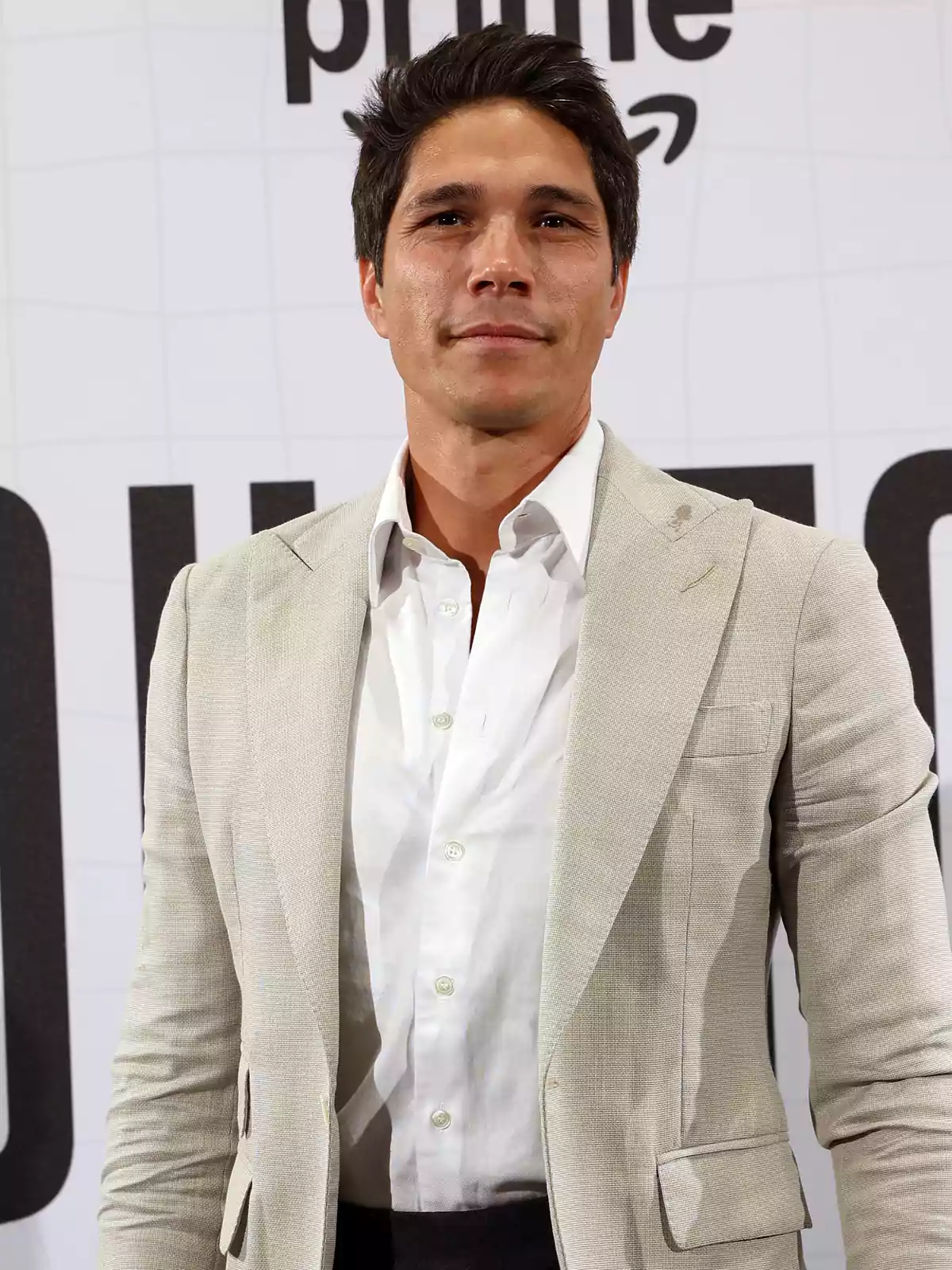 Hombre de cabello oscuro con saco beige y camisa blanca posando frente a un fondo con letras grandes. Hombre de cabello oscuro con saco beige y camisa blanca posando frente a un fondo con letras grandes.