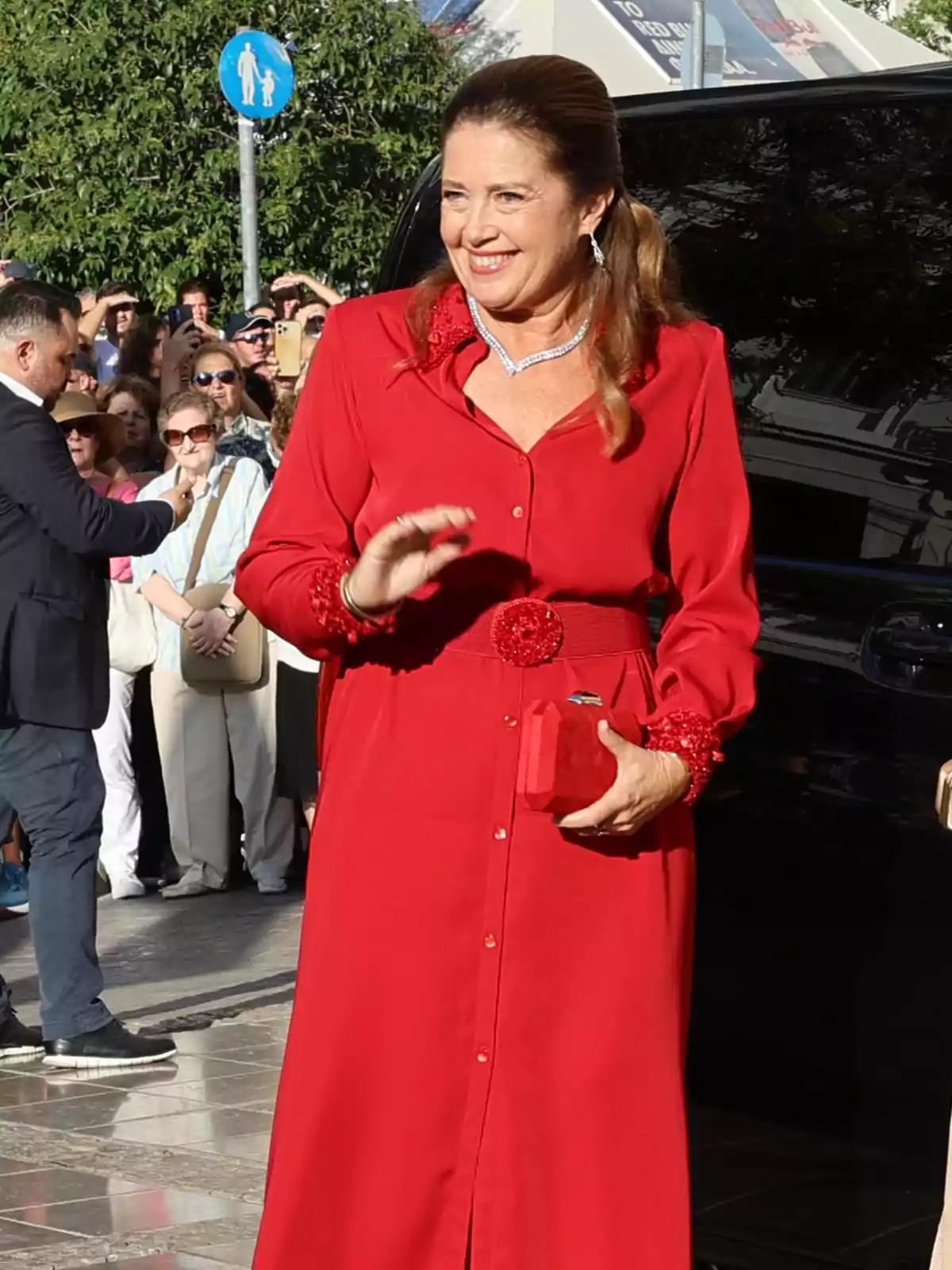 Mujer sonriente con vestido rojo largo y bolso a juego saludando mientras camina por la calle rodeada de gente Mujer sonriente con vestido rojo largo y bolso a juego saludando mientras camina por la calle rodeada de gente