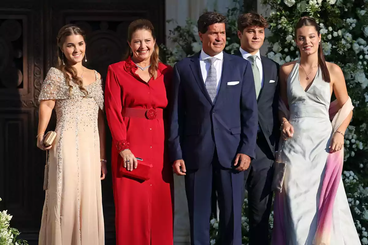 Cinco personas elegantemente vestidas posan juntas al aire libre frente a una puerta decorada y rodeada de flores blancas Cinco personas elegantemente vestidas posan juntas al aire libre frente a una puerta decorada y rodeada de flores blancas