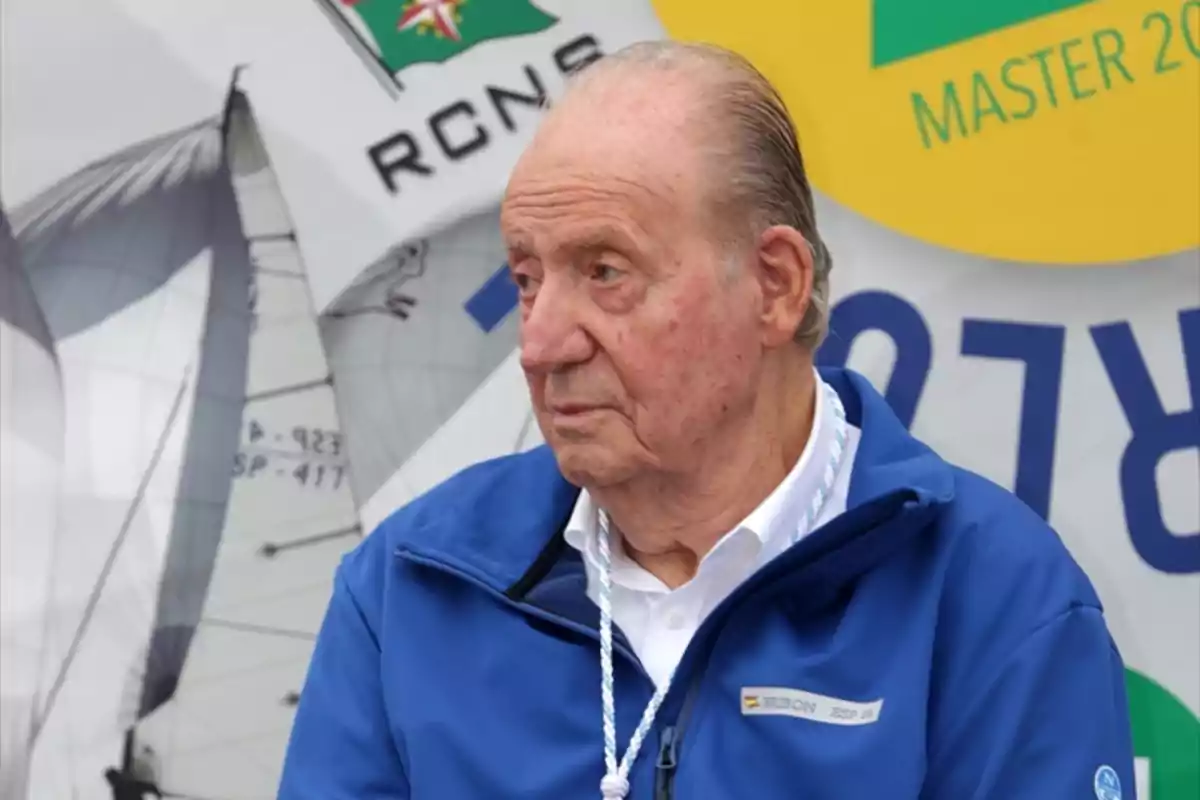 Hombre mayor con chaqueta azul sentado frente a un fondo con velas de barco y logotipos náuticos Hombre mayor con chaqueta azul sentado frente a un fondo con velas de barco y logotipos náuticos