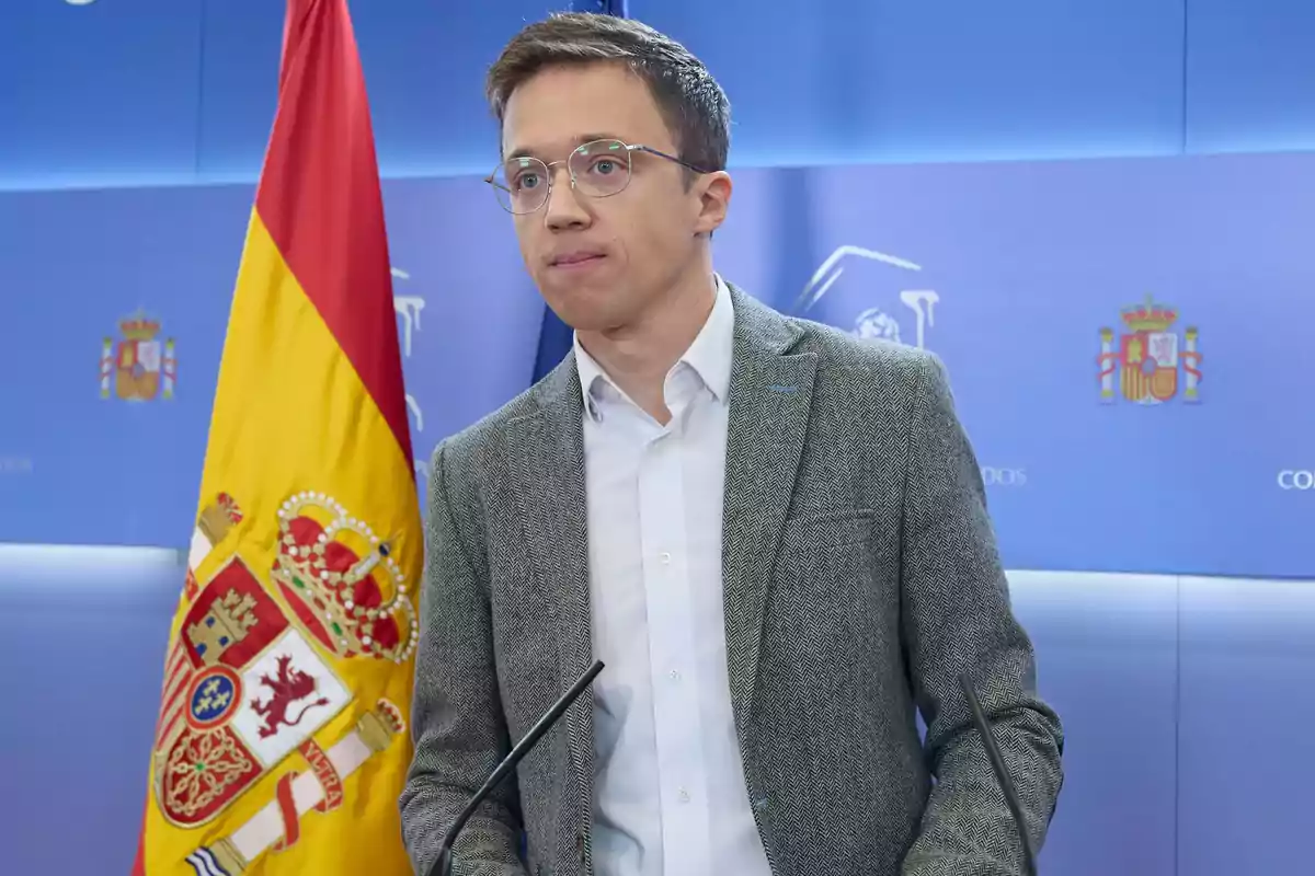 Hombre con gafas y chaqueta gris hablando en una rueda de prensa con una bandera de España al fondo Hombre con gafas y chaqueta gris hablando en una rueda de prensa con una bandera de España al fondo