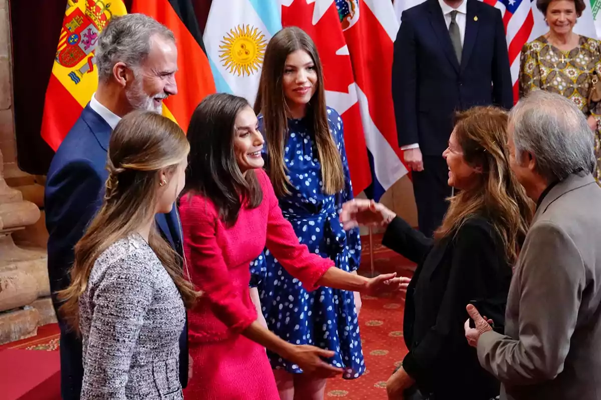 Un grupo de personas elegantemente vestidas se saluda y sonríe en un evento formal con varias banderas de diferentes países al fondo Un grupo de personas elegantemente vestidas se saluda y sonríe en un evento formal con varias banderas de diferentes países al fondo