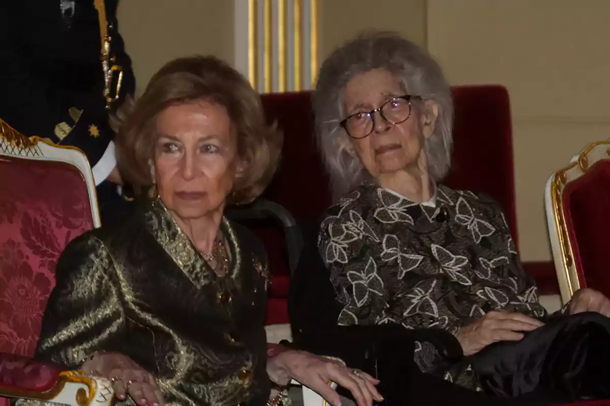 Dos mujeres mayores elegantemente vestidas están sentadas en sillas tapizadas de rojo en un ambiente formal Dos mujeres mayores elegantemente vestidas están sentadas en sillas tapizadas de rojo en un ambiente formal