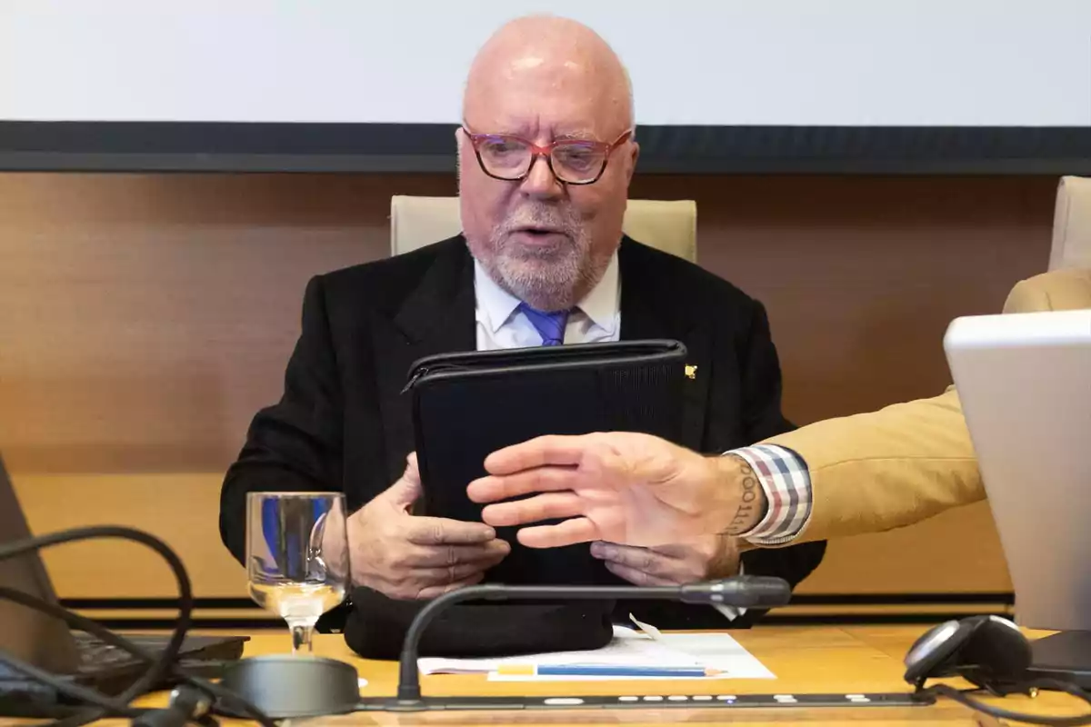 Hombre mayor con gafas y barba sentado en una mesa de conferencia sosteniendo una carpeta negra mientras una mano se interpone frente a él Hombre mayor con gafas y barba sentado en una mesa de conferencia sosteniendo una carpeta negra mientras una mano se interpone frente a él