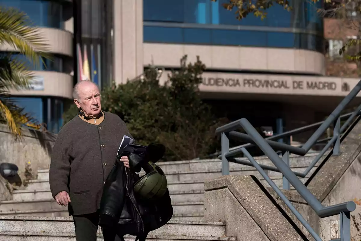 Un hombre mayor camina por unas escaleras al aire libre frente a un edificio con el letrero Audiencia Provincial de Madrid mientras sostiene una chaqueta y un casco en la mano. Un hombre mayor camina por unas escaleras al aire libre frente a un edificio con el letrero Audiencia Provincial de Madrid mientras sostiene una chaqueta y un casco en la mano.