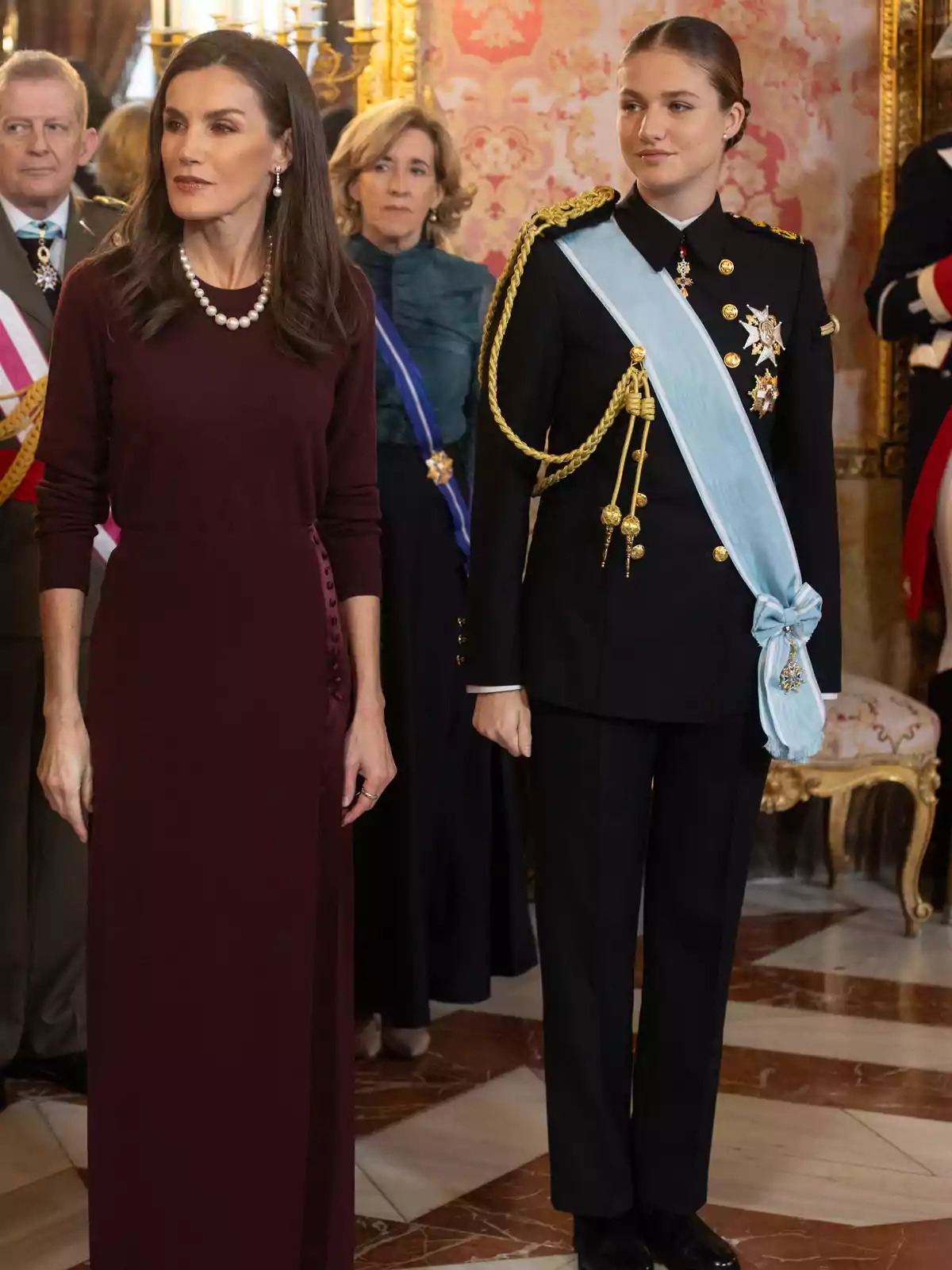 Dos mujeres en un evento formal, una con vestido burdeos y collar de perlas, la otra con uniforme militar y banda azul, rodeadas de otras personas en un elegante salón decorado. Dos mujeres en un evento formal, una con vestido burdeos y collar de perlas, la otra con uniforme militar y banda azul, rodeadas de otras personas en un elegante salón decorado.