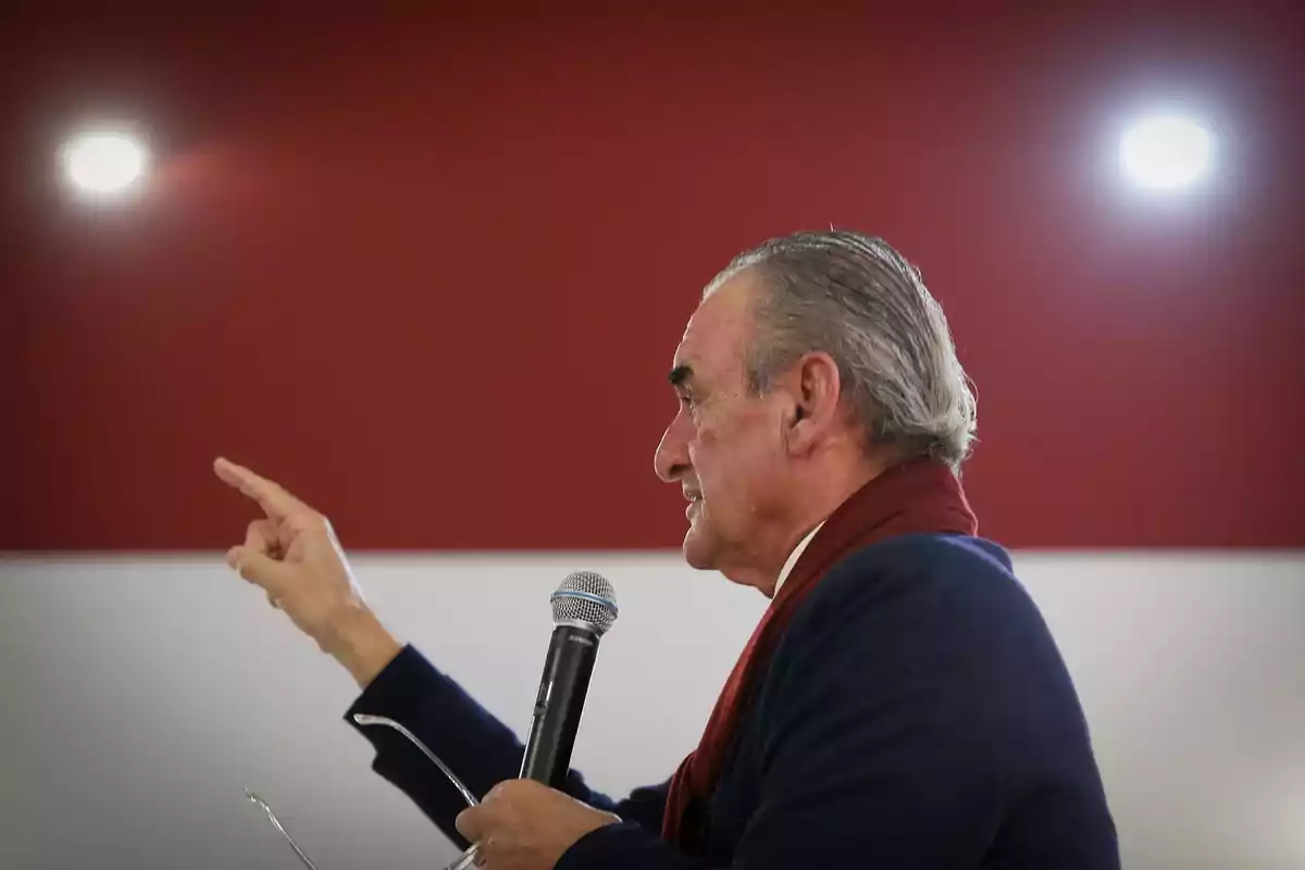 Hombre mayor de perfil hablando por micrófono y señalando con el dedo en un fondo rojo y blanco Hombre mayor de perfil hablando por micrófono y señalando con el dedo en un fondo rojo y blanco