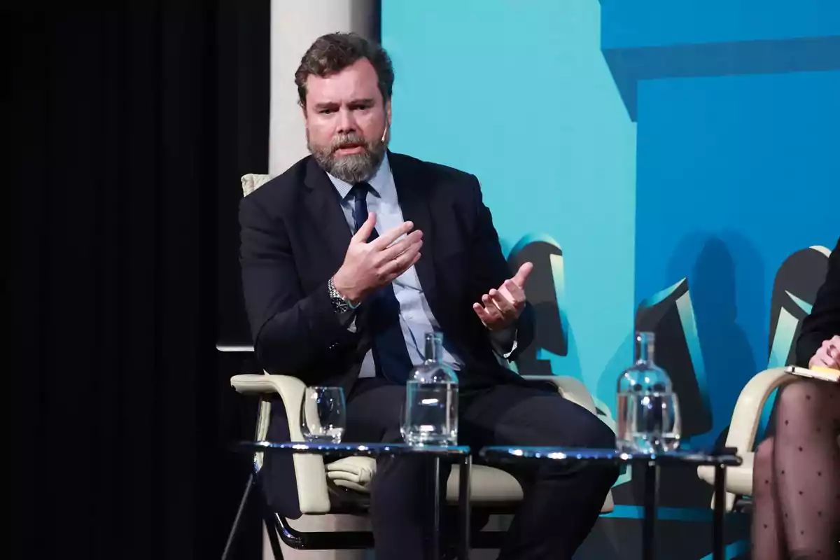 Hombre con barba y traje oscuro sentado en una silla participando en una conversación en un evento profesional con fondo azul y mesa de cristal con botellas de agua al frente Hombre con barba y traje oscuro sentado en una silla participando en una conversación en un evento profesional con fondo azul y mesa de cristal con botellas de agua al frente