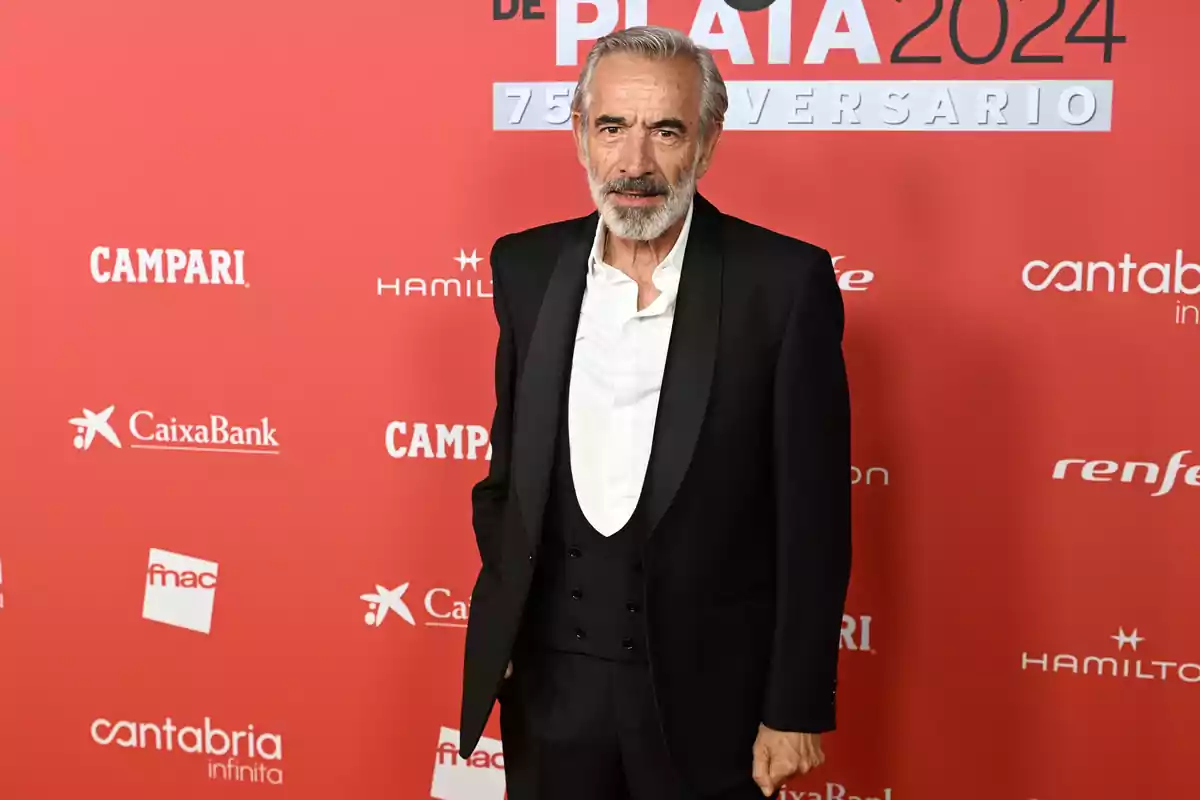 Hombre de cabello y barba canosos con traje negro y camisa blanca posando frente a un fondo rojo con logotipos de marcas y texto de un evento de 2024 Hombre de cabello y barba canosos con traje negro y camisa blanca posando frente a un fondo rojo con logotipos de marcas y texto de un evento de 2024