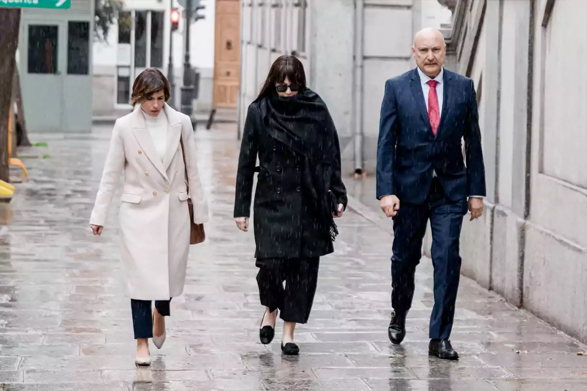 Tres personas caminan bajo la lluvia por una acera mojada, una mujer con abrigo claro, otra con abrigo oscuro y gafas de sol, y un hombre con traje azul y corbata roja. Tres personas caminan bajo la lluvia por una acera mojada, una mujer con abrigo claro, otra con abrigo oscuro y gafas de sol, y un hombre con traje azul y corbata roja.