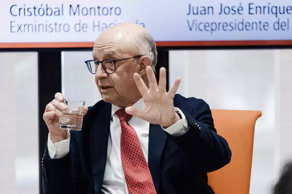 Hombre mayor con gafas y corbata roja sentado en una silla naranja, sosteniendo un vaso de agua y gesticulando con la mano mientras habla en un evento Hombre mayor con gafas y corbata roja sentado en una silla naranja, sosteniendo un vaso de agua y gesticulando con la mano mientras habla en un evento