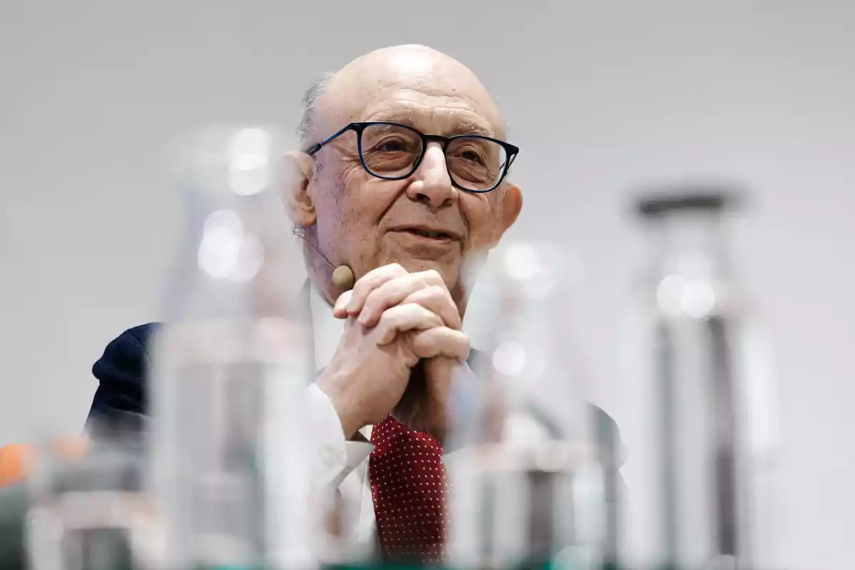 Hombre mayor con gafas y micrófono de diadema hablando en un evento, con botellas desenfocadas en primer plano Hombre mayor con gafas y micrófono de diadema hablando en un evento, con botellas desenfocadas en primer plano