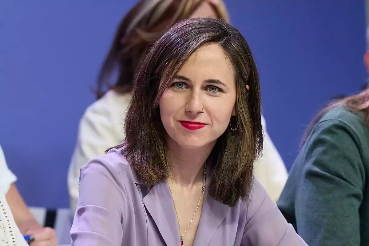 Mujer de cabello castaño y labios rojos sentada en una mesa con fondo azul