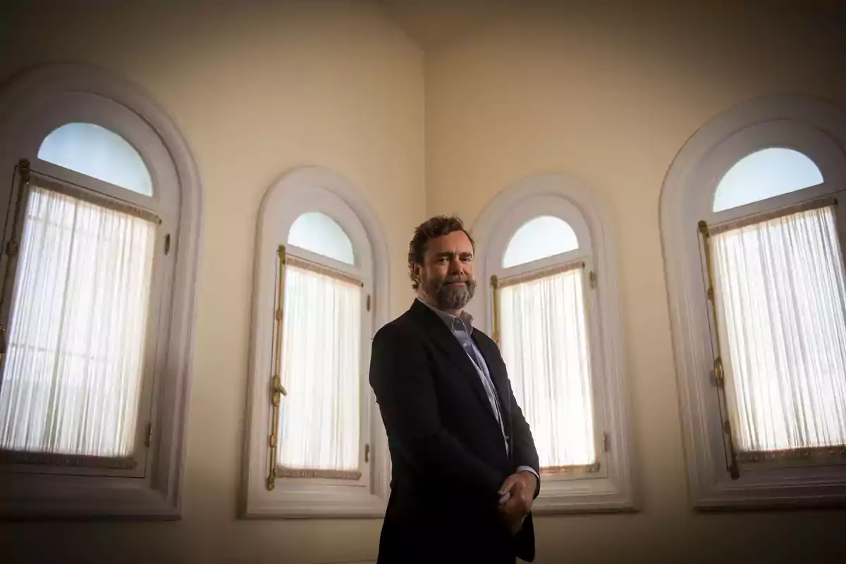 Hombre de barba y traje oscuro posando en una habitación iluminada con cuatro ventanas altas de marco blanco y cortinas translúcidas. Hombre de barba y traje oscuro posando en una habitación iluminada con cuatro ventanas altas de marco blanco y cortinas translúcidas.