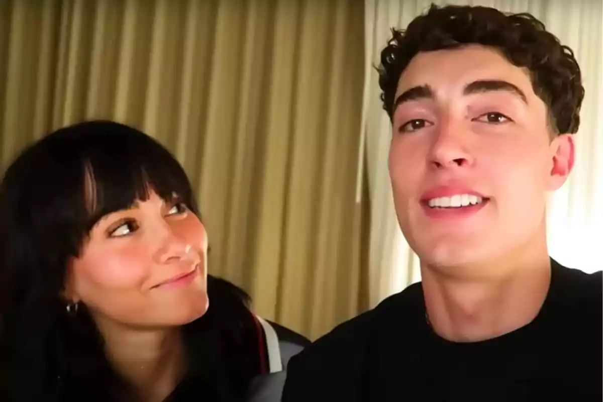 Una mujer de cabello oscuro mira sonriente a un hombre de cabello rizado que está sonriendo frente a la cámara, ambos en un ambiente interior con cortinas de fondo. Una mujer de cabello oscuro mira sonriente a un hombre de cabello rizado que está sonriendo frente a la cámara, ambos en un ambiente interior con cortinas de fondo.