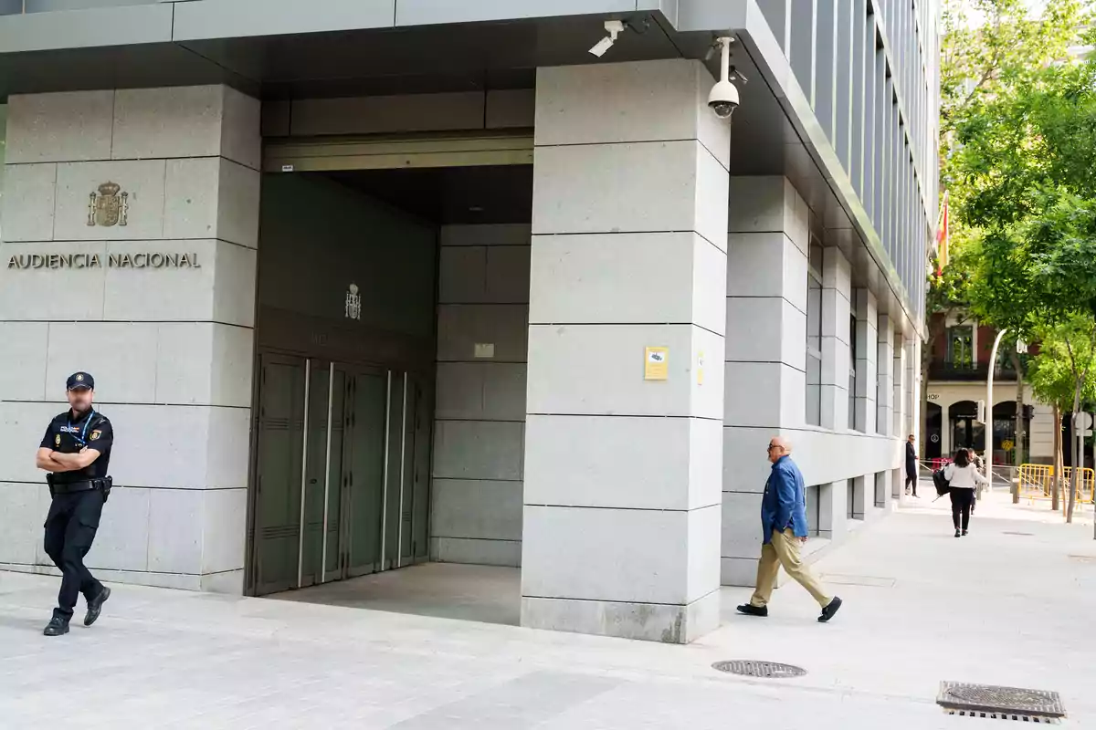 Fachada de un edificio gubernamental con un policía de pie en la entrada y varias personas caminando por la acera Fachada de un edificio gubernamental con un policía de pie en la entrada y varias personas caminando por la acera