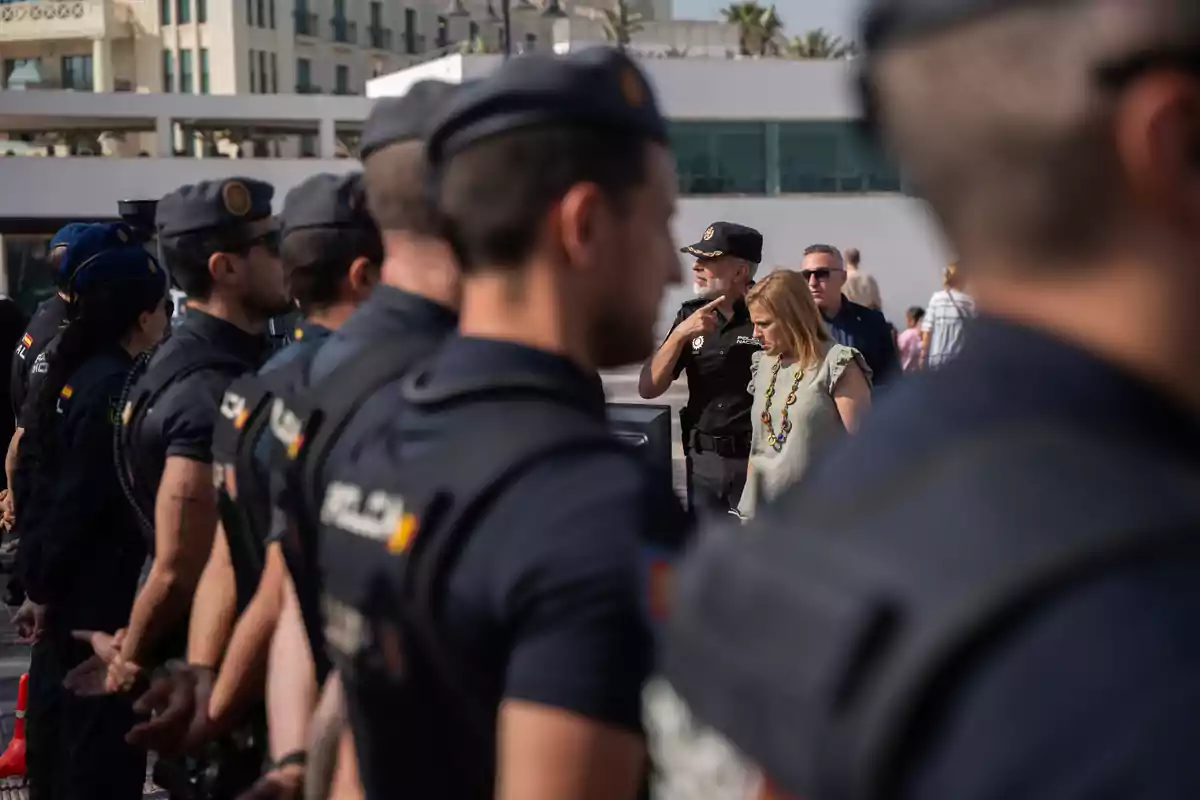 Varios policías uniformados forman una fila mientras una mujer y un oficial conversan al fondo en un entorno urbano. Varios policías uniformados forman una fila mientras una mujer y un oficial conversan al fondo en un entorno urbano.