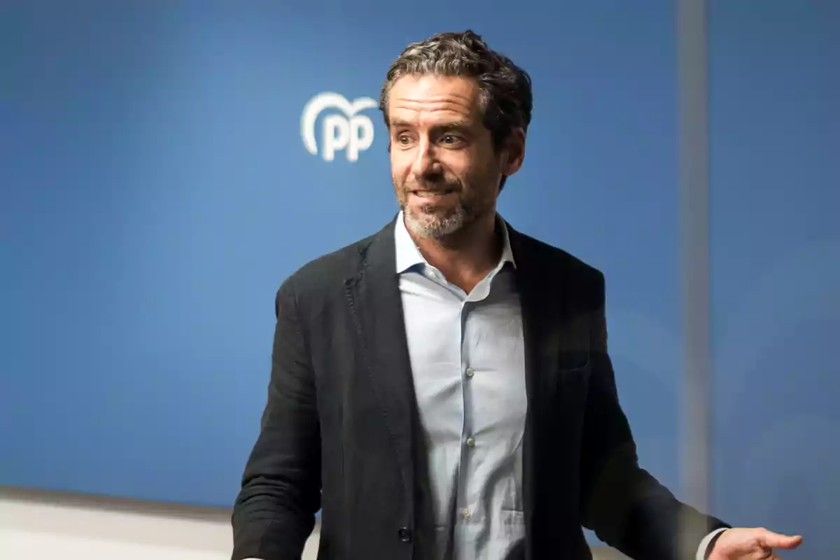 Hombre de cabello rizado y barba con chaqueta oscura y camisa clara de pie frente a una pared azul con el logotipo de un partido político al fondo Hombre de cabello rizado y barba con chaqueta oscura y camisa clara de pie frente a una pared azul con el logotipo de un partido político al fondo