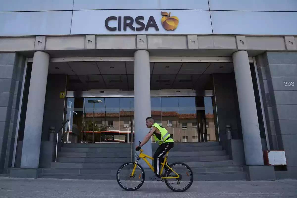 Un hombre con chaleco reflectante monta una bicicleta amarilla frente a la entrada de un edificio con el letrero CIRSA.