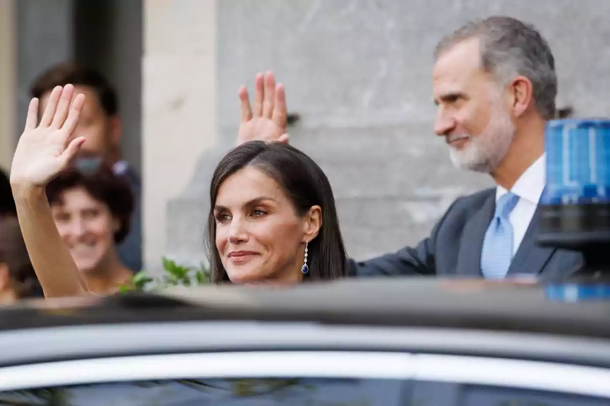 Una mujer y un hombre elegantemente vestidos saludan con la mano mientras están de pie junto a un coche en un evento público Una mujer y un hombre elegantemente vestidos saludan con la mano mientras están de pie junto a un coche en un evento público