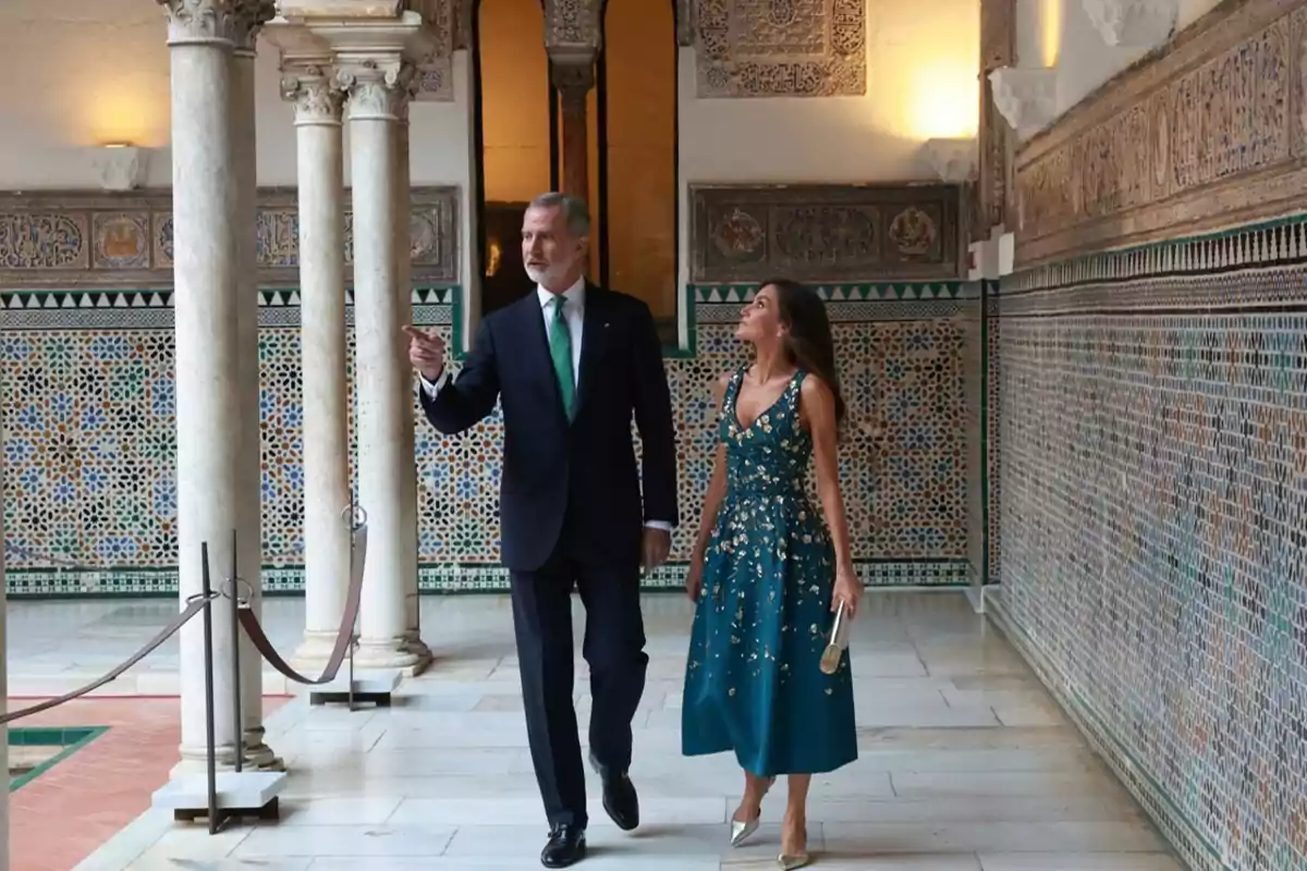 Un hombre con traje oscuro y corbata verde camina junto a una mujer con vestido azul y detalles dorados en un pasillo decorado con mosaicos y columnas de estilo árabe Un hombre con traje oscuro y corbata verde camina junto a una mujer con vestido azul y detalles dorados en un pasillo decorado con mosaicos y columnas de estilo árabe