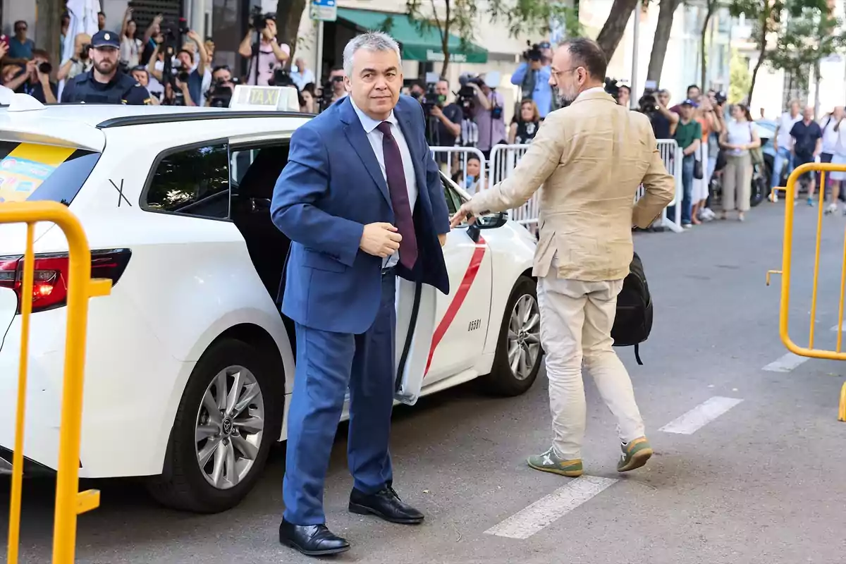 Un hombre con traje azul sale de un taxi blanco mientras otra persona con chaqueta beige y mochila lo acompaña, al fondo hay periodistas y cámaras detrás de vallas en una calle urbana. Un hombre con traje azul sale de un taxi blanco mientras otra persona con chaqueta beige y mochila lo acompaña, al fondo hay periodistas y cámaras detrás de vallas en una calle urbana.