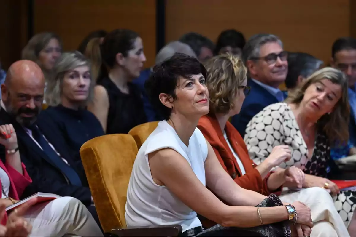 Mujer de cabello corto y oscuro sentada en una sala de conferencias rodeada de otras personas que también están atentas al evento