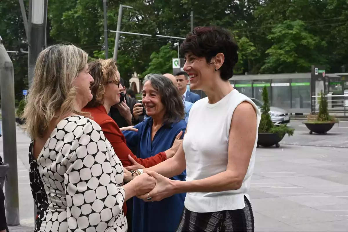 Varias mujeres sonríen y se saludan cordialmente al aire libre en una zona urbana rodeada de árboles y paradas de transporte público. Varias mujeres sonríen y se saludan cordialmente al aire libre en una zona urbana rodeada de árboles y paradas de transporte público.