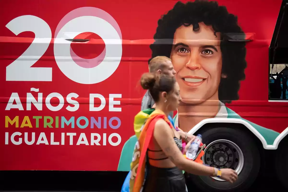 Personas caminando frente a un autobús rojo decorado con un retrato y un mensaje que celebra 20 años de matrimonio igualitario Personas caminando frente a un autobús rojo decorado con un retrato y un mensaje que celebra 20 años de matrimonio igualitario