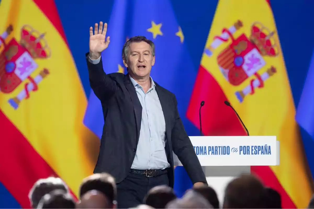 Hombre de traje saludando con la mano levantada en un mitin político frente a banderas de España y la Unión Europea Hombre de traje saludando con la mano levantada en un mitin político frente a banderas de España y la Unión Europea