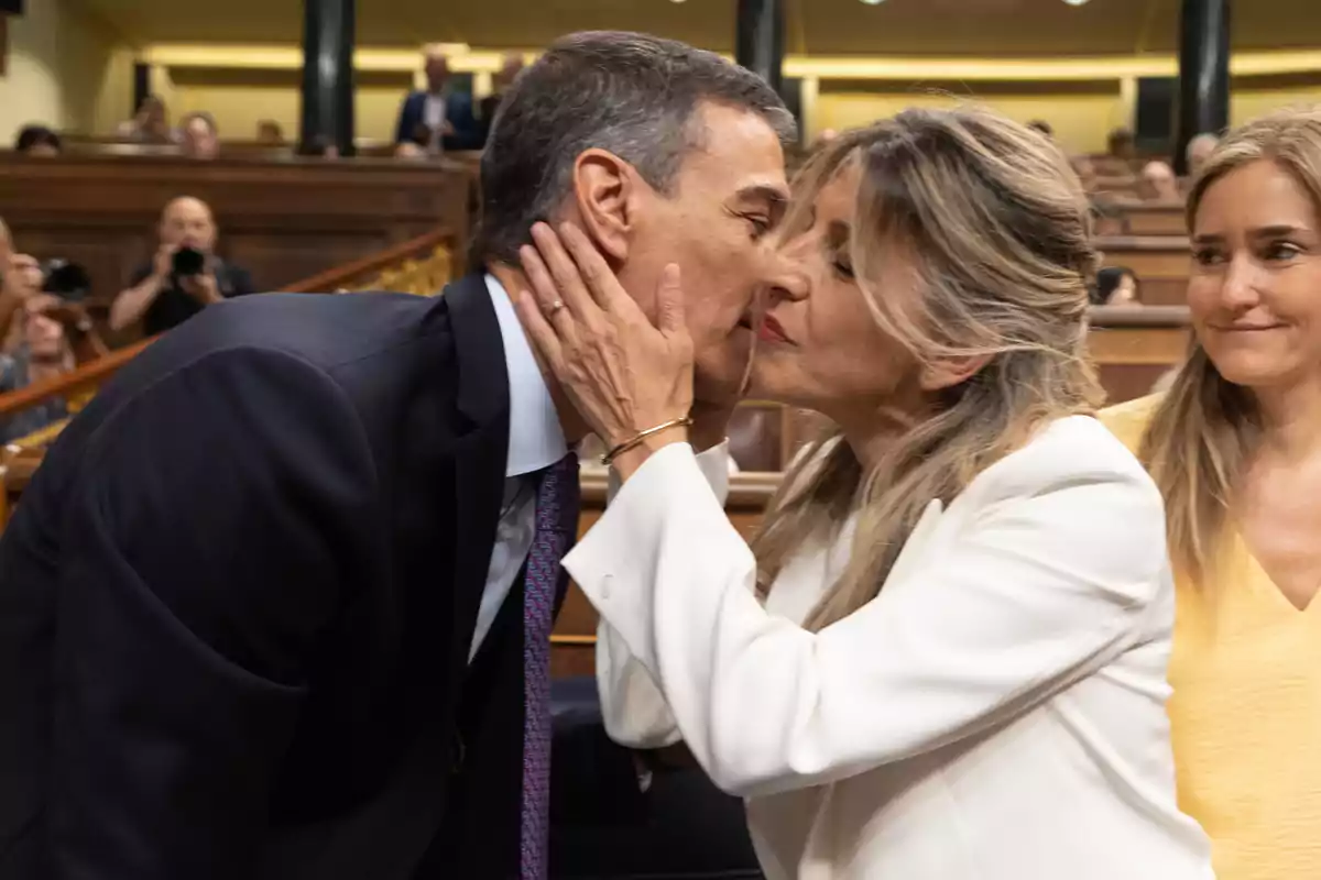 Un hombre y una mujer se saludan con un beso en la mejilla en un entorno formal mientras otra mujer observa sonriente Un hombre y una mujer se saludan con un beso en la mejilla en un entorno formal mientras otra mujer observa sonriente