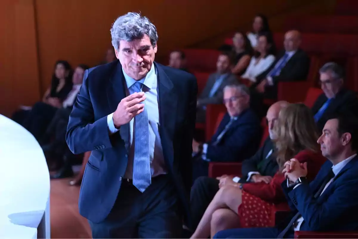 Hombre de cabello canoso con traje y corbata caminando en un auditorio mientras varias personas sentadas observan desde el fondo Hombre de cabello canoso con traje y corbata caminando en un auditorio mientras varias personas sentadas observan desde el fondo