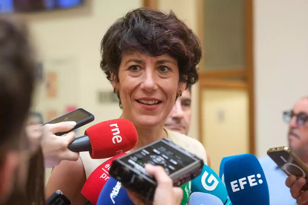 Mujer de cabello corto y oscuro sonriendo mientras es entrevistada por varios periodistas que sostienen micrófonos de diferentes medios de comunicación Mujer de cabello corto y oscuro sonriendo mientras es entrevistada por varios periodistas que sostienen micrófonos de diferentes medios de comunicación