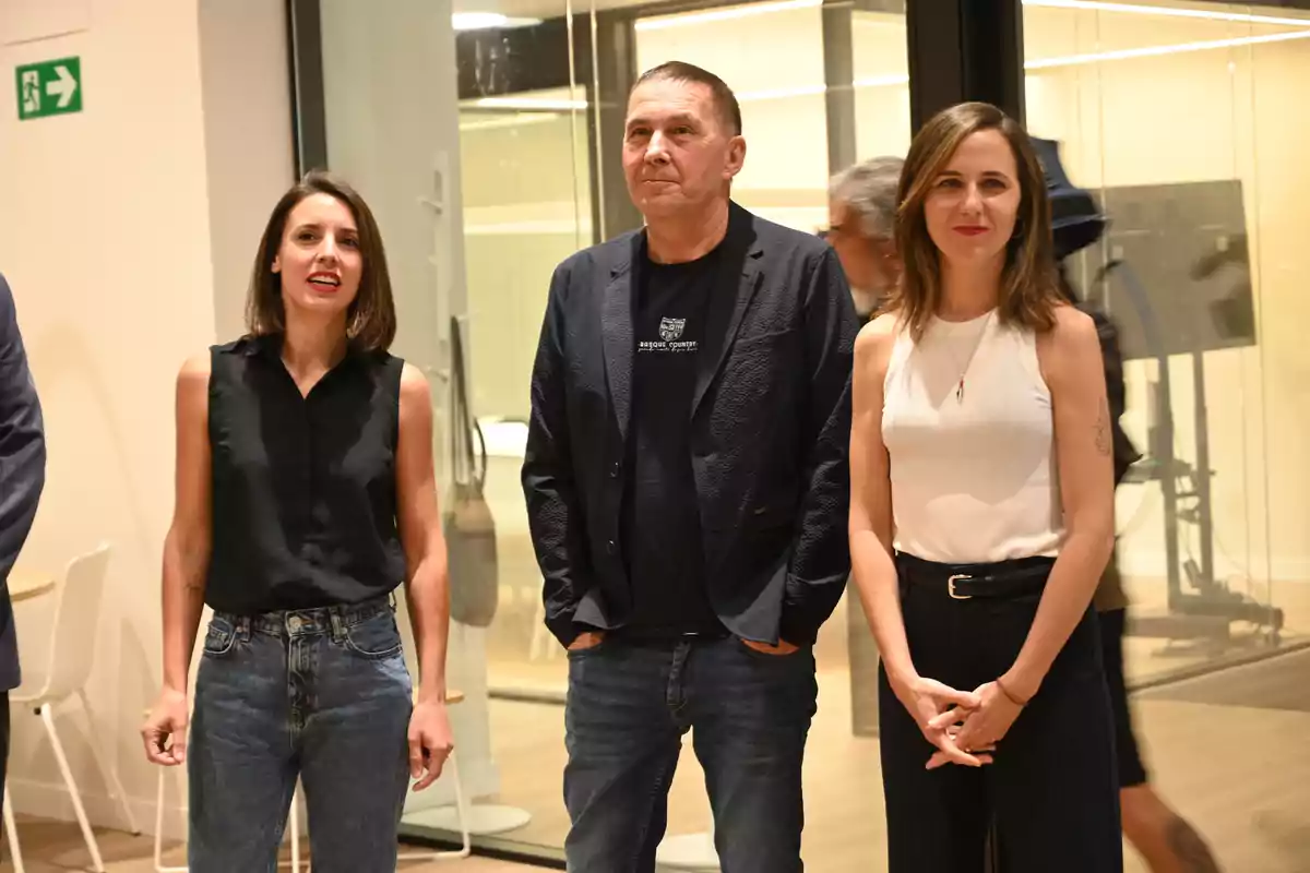 Tres personas posan de pie en un espacio interior moderno con paredes de cristal y mobiliario minimalista, dos mujeres a los lados y un hombre en el centro, todos vestidos de manera casual elegante. Tres personas posan de pie en un espacio interior moderno con paredes de cristal y mobiliario minimalista, dos mujeres a los lados y un hombre en el centro, todos vestidos de manera casual elegante.