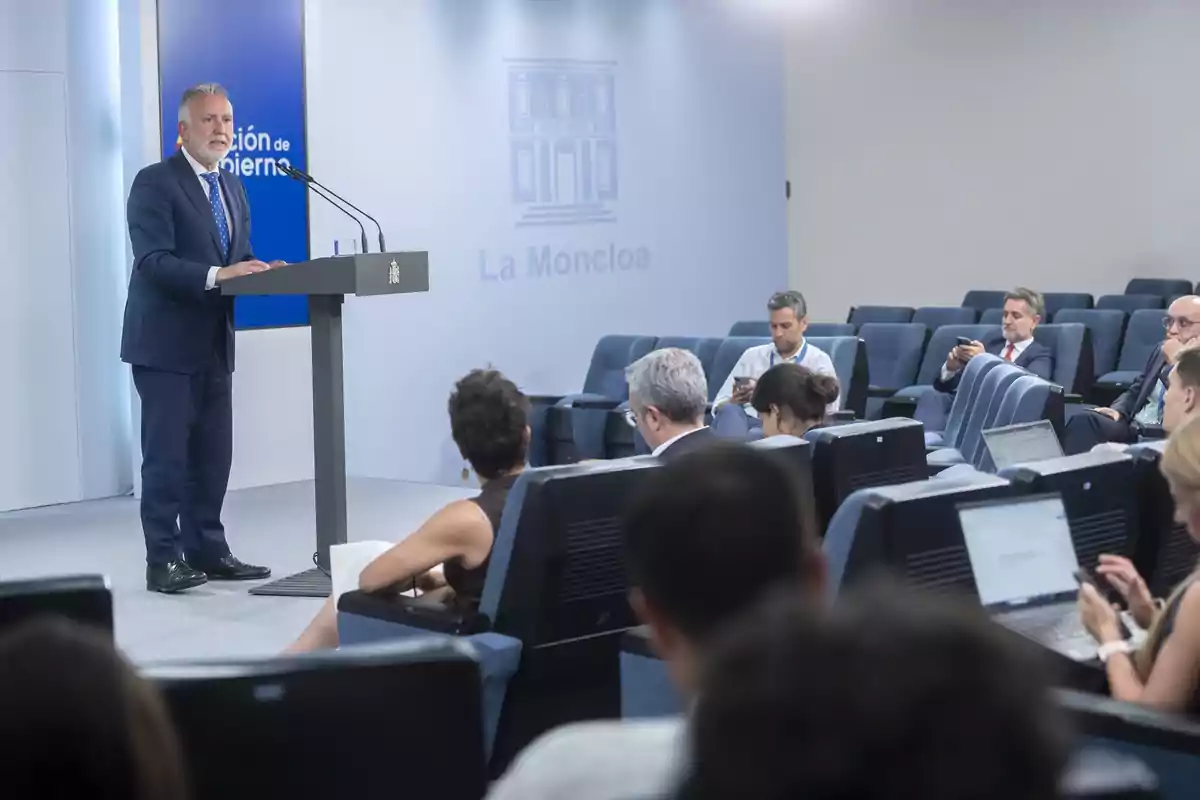 Un hombre de traje habla en un atril frente a varias personas sentadas en una sala de conferencias, algunas tomando notas en computadoras portátiles y otras usando sus teléfonos móviles. Un hombre de traje habla en un atril frente a varias personas sentadas en una sala de conferencias, algunas tomando notas en computadoras portátiles y otras usando sus teléfonos móviles.