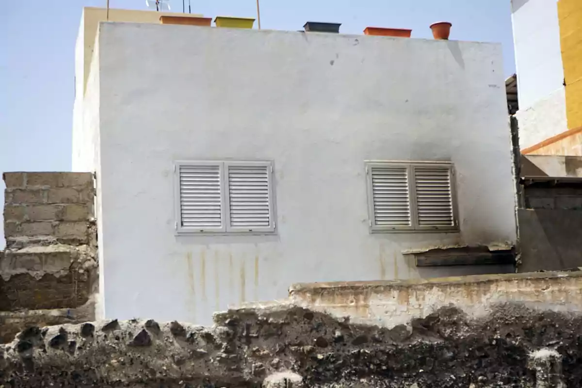 Casa blanca con dos ventanas pequeñas y persianas cerradas, macetas de colores en el techo y una pared de piedra en primer plano