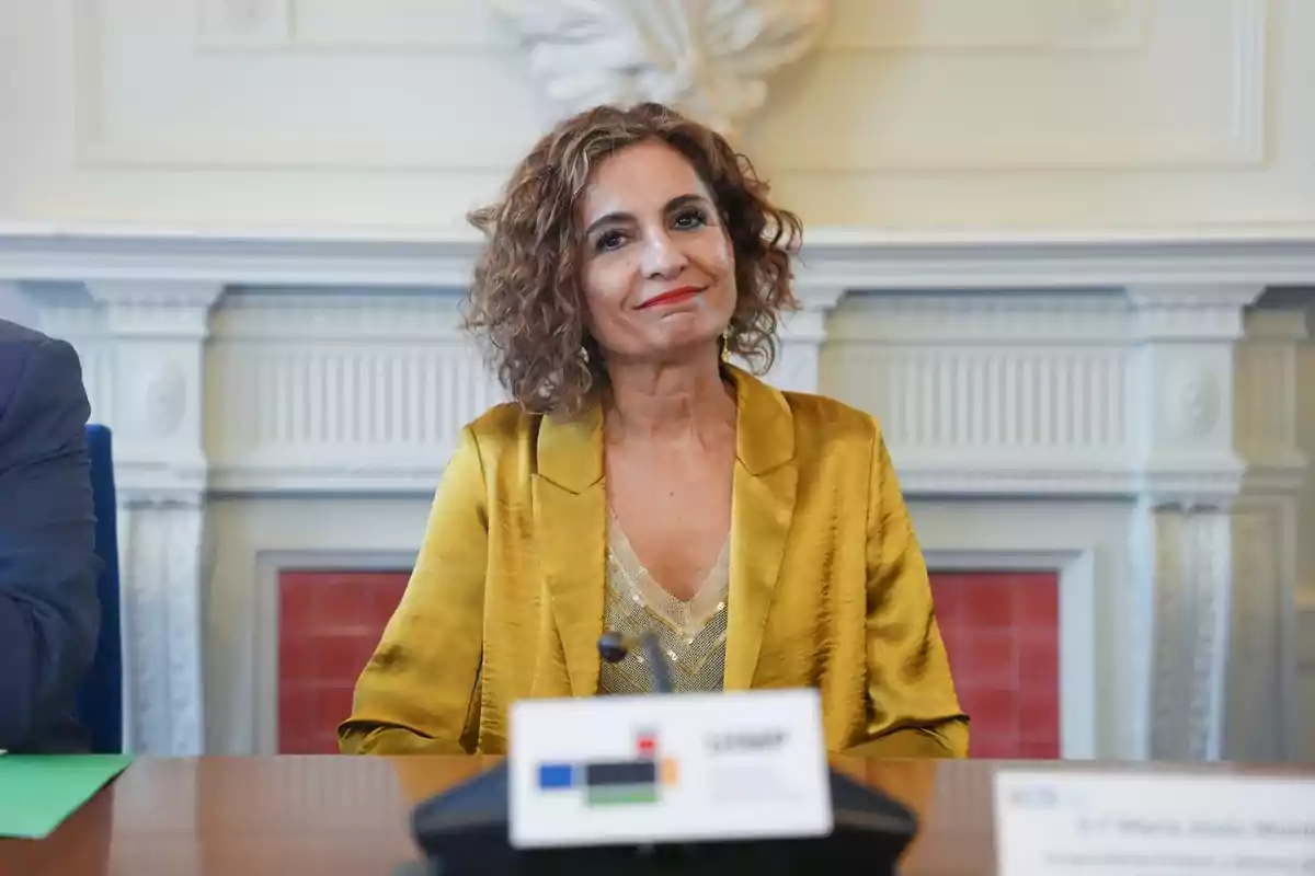 Mujer de cabello rizado y corto con chaqueta dorada sentada en una mesa durante una reunión en una sala elegante Mujer de cabello rizado y corto con chaqueta dorada sentada en una mesa durante una reunión en una sala elegante