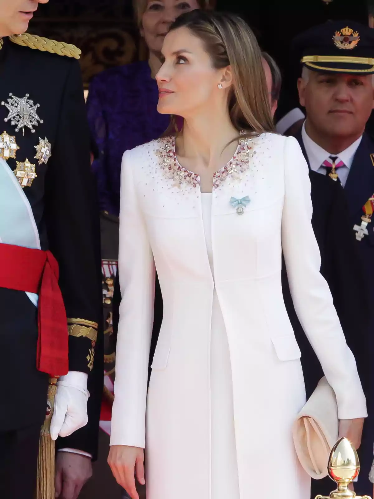 Mujer con abrigo blanco elegante decorado con pedrería en el cuello, sosteniendo un bolso beige y mirando hacia un hombre uniformado en un evento formal Mujer con abrigo blanco elegante decorado con pedrería en el cuello, sosteniendo un bolso beige y mirando hacia un hombre uniformado en un evento formal