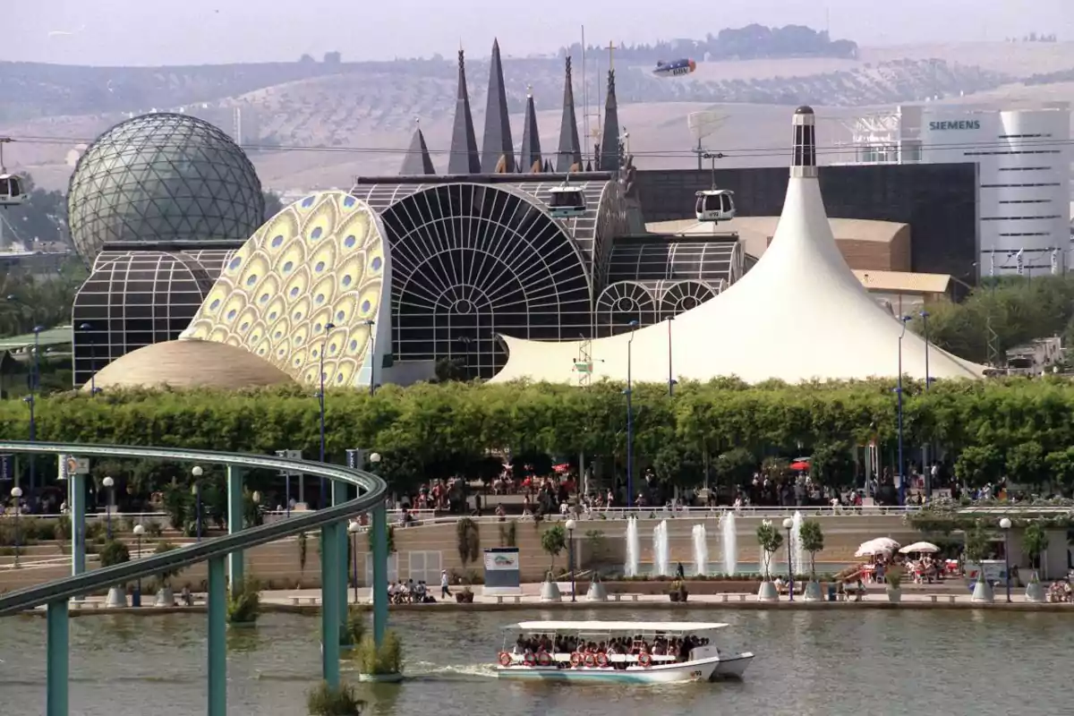 Edificios de arquitectura futurista y colorida junto a un lago con una embarcación llena de personas y un monorraíl elevado rodeados de árboles y fuentes Edificios de arquitectura futurista y colorida junto a un lago con una embarcación llena de personas y un monorraíl elevado rodeados de árboles y fuentes