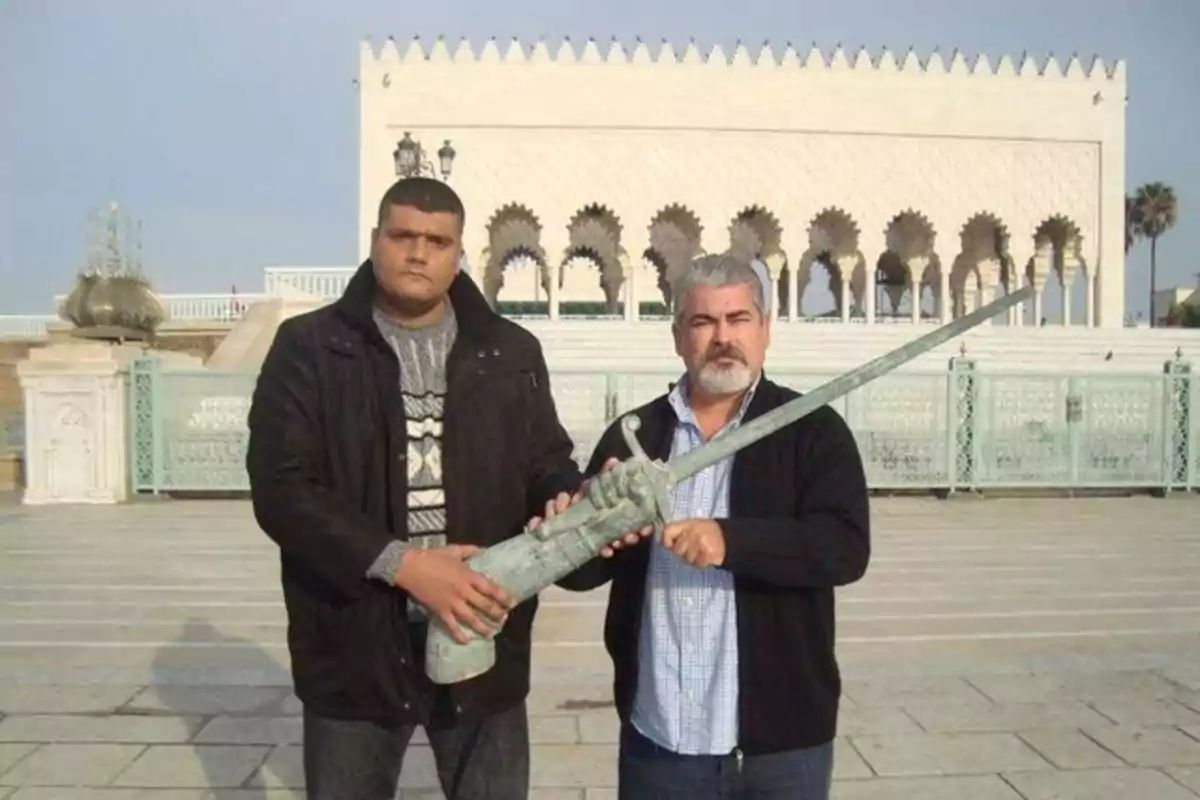 Dos hombres posan al aire libre sosteniendo juntos una gran espada de metal frente a un edificio de arquitectura árabe Dos hombres posan al aire libre sosteniendo juntos una gran espada de metal frente a un edificio de arquitectura árabe