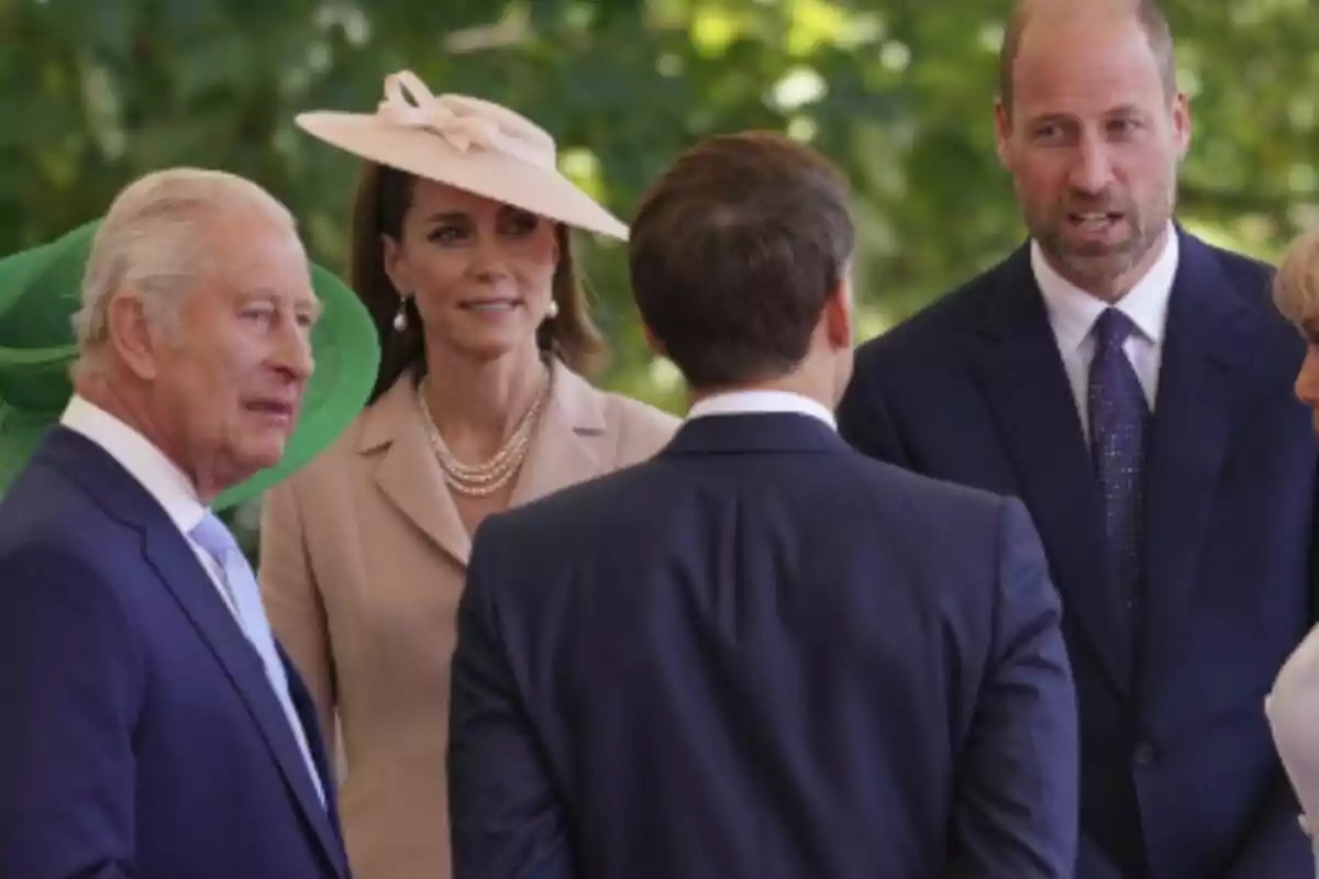 Varias personas elegantemente vestidas conversan al aire libre en un evento formal, las cuales son la Familia Real británica junto a Emmanuel Macron.