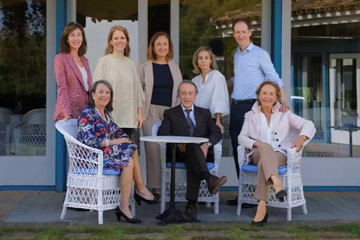 Ocho personas posan sonrientes frente a una casa con grandes ventanales, tres de ellas están sentadas en sillas de mimbre blanco alrededor de una mesa pequeña y las otras cinco están de pie detrás de ellas.