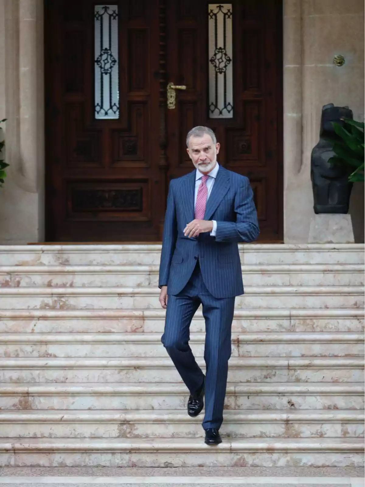 Hombre de cabello canoso con barba corta, vestido con traje azul de rayas y corbata rosa, bajando unas escaleras de mármol frente a una puerta de madera tallada Hombre de cabello canoso con barba corta, vestido con traje azul de rayas y corbata rosa, bajando unas escaleras de mármol frente a una puerta de madera tallada