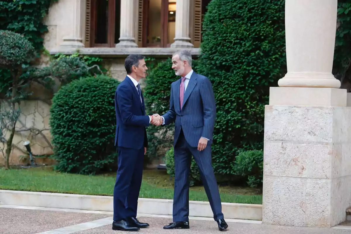 Dos hombres vestidos con traje se dan la mano en un jardín elegante frente a un edificio de piedra con columnas y arbustos recortados Dos hombres vestidos con traje se dan la mano en un jardín elegante frente a un edificio de piedra con columnas y arbustos recortados