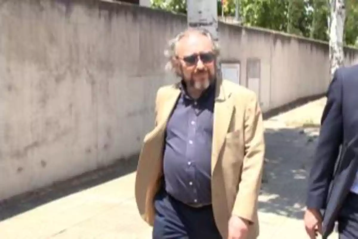 Hombre con gafas de sol y chaqueta beige caminando por una acera junto a una pared bajo la luz del sol Hombre con gafas de sol y chaqueta beige caminando por una acera junto a una pared bajo la luz del sol